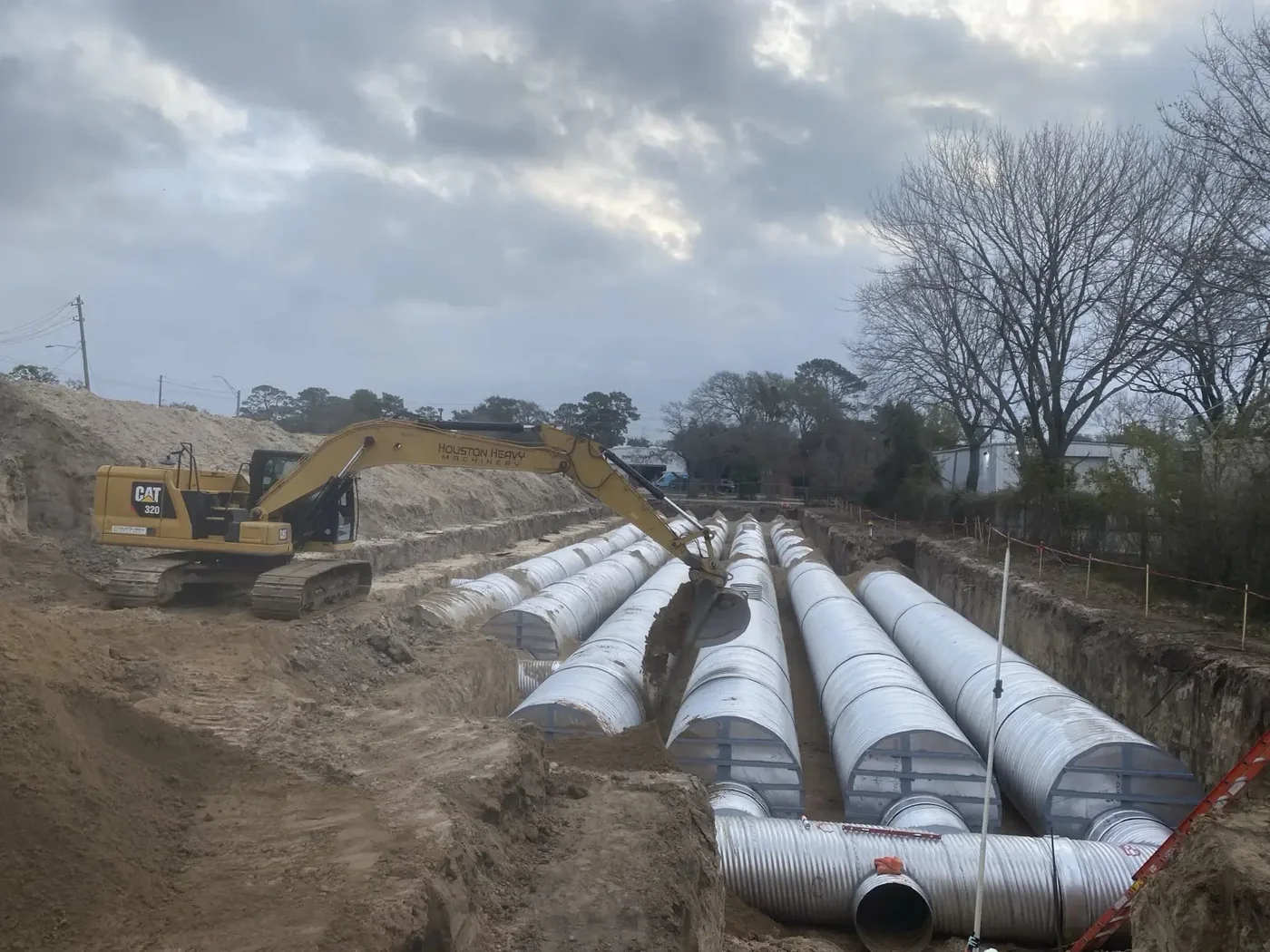 Tracked excavator digging above rows of large corrugated metal underground detention pipe in a deep commercial jobsite trench