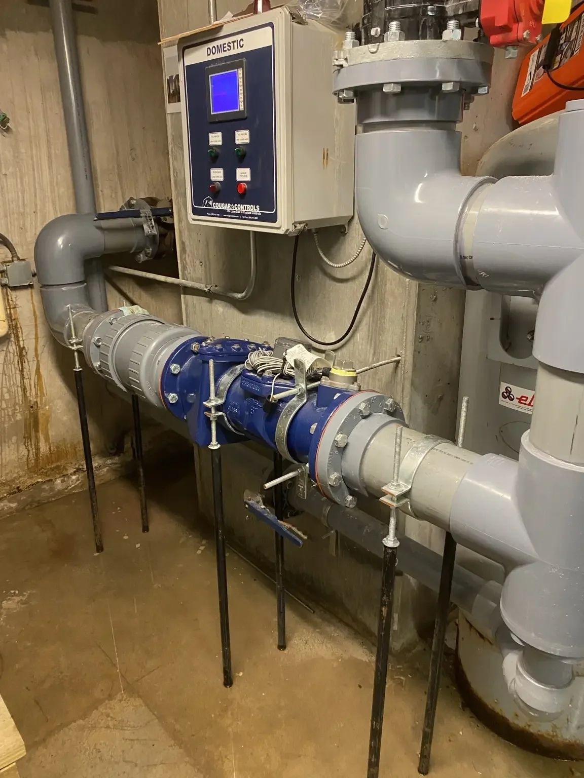 Interior domestic water service with grey piping, a blue backflow valve assembly and a wall-mounted control panel