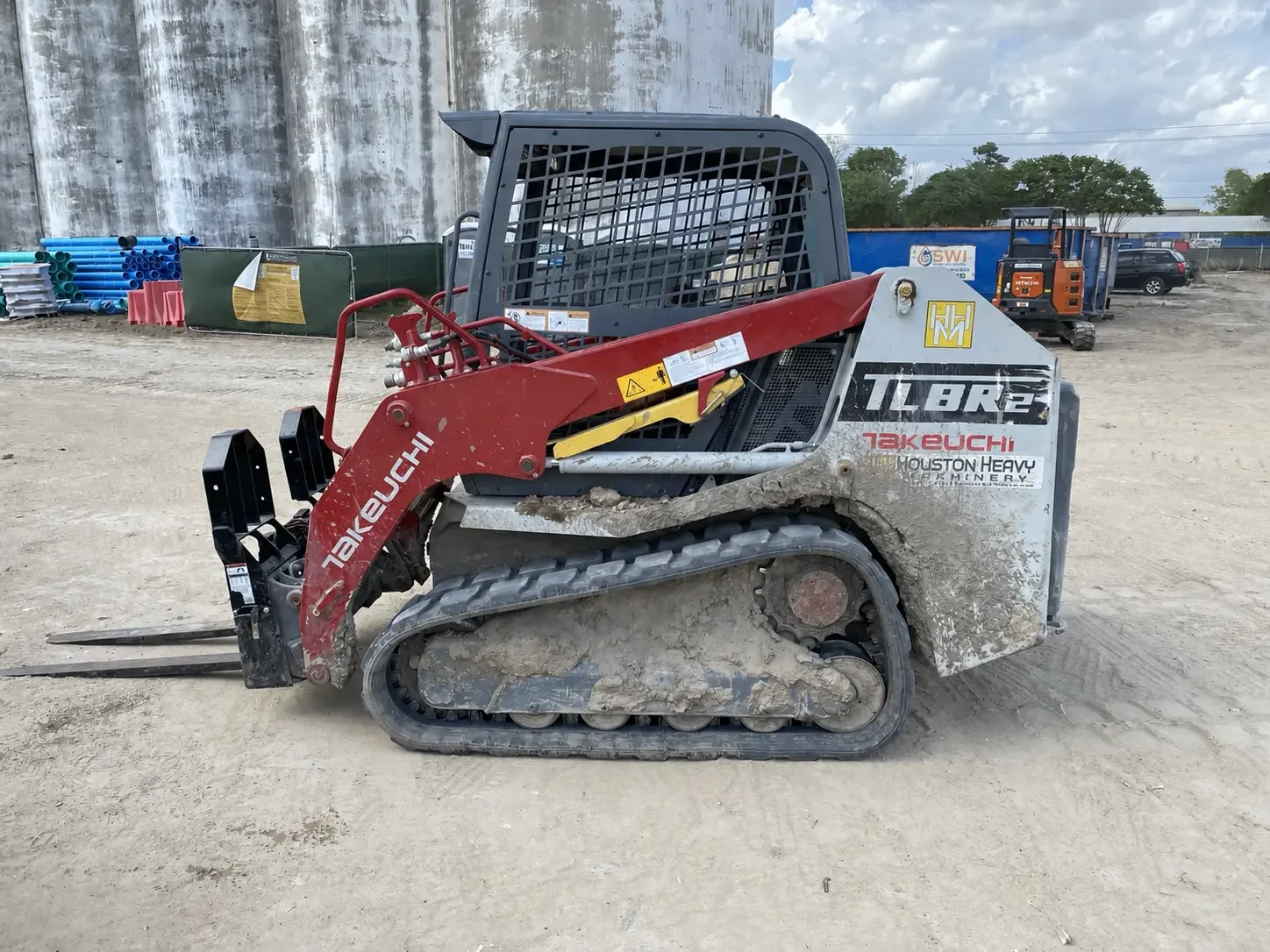 Compact track loader fitted with pallet forks parked on a laydown yard beside stacked pipe