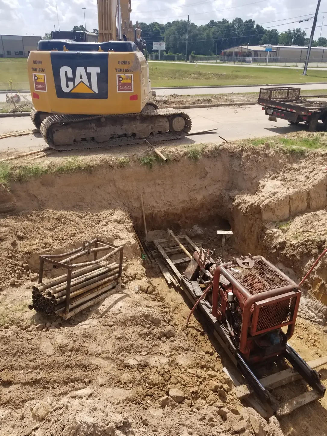 Auger boring rig set on rails inside a bore pit with drill rods staged nearby and an excavator above the pit