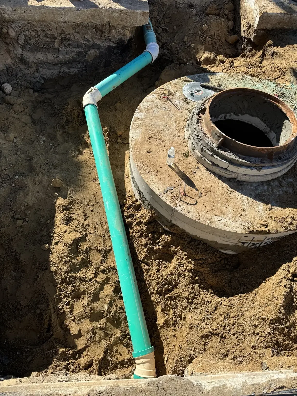 Top-down view of a concrete manhole base with green PVC sewer lines entering at angles in a sandy trench