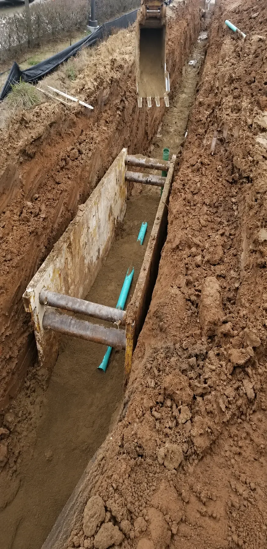 Deep vertical utility trench protected by steel trench shoring boxes with green PVC pipe at the bottom