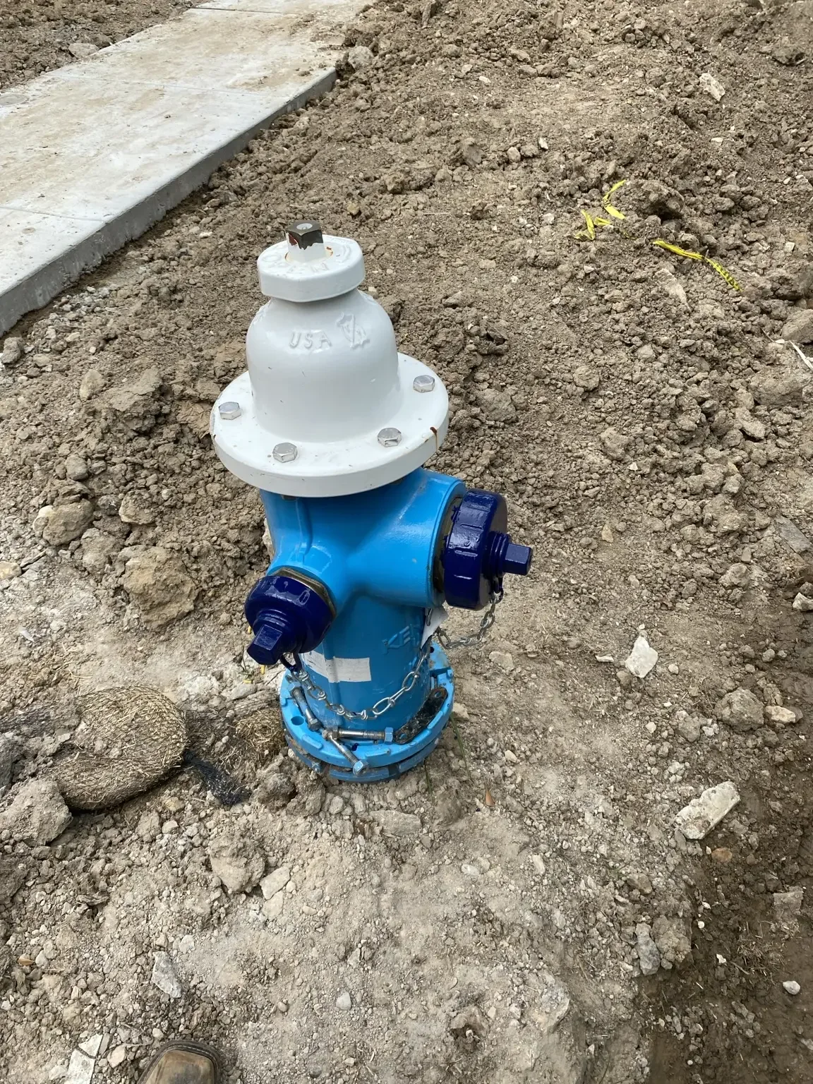 Newly installed blue and white fire hydrant set in graded dirt next to a concrete sidewalk