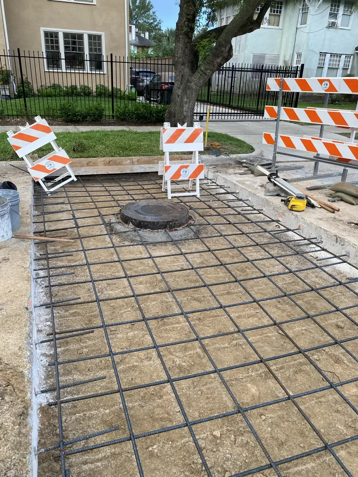 Rebar grid and edge forms set around an existing manhole cover ahead of a concrete pour, closed off with barricades