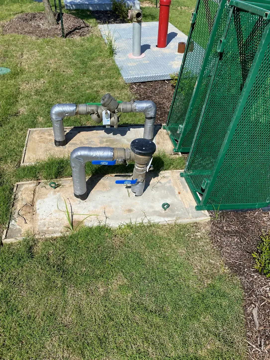 Two above-grade backflow prevention assemblies mounted on concrete pads near a fire hydrant and protective enclosure