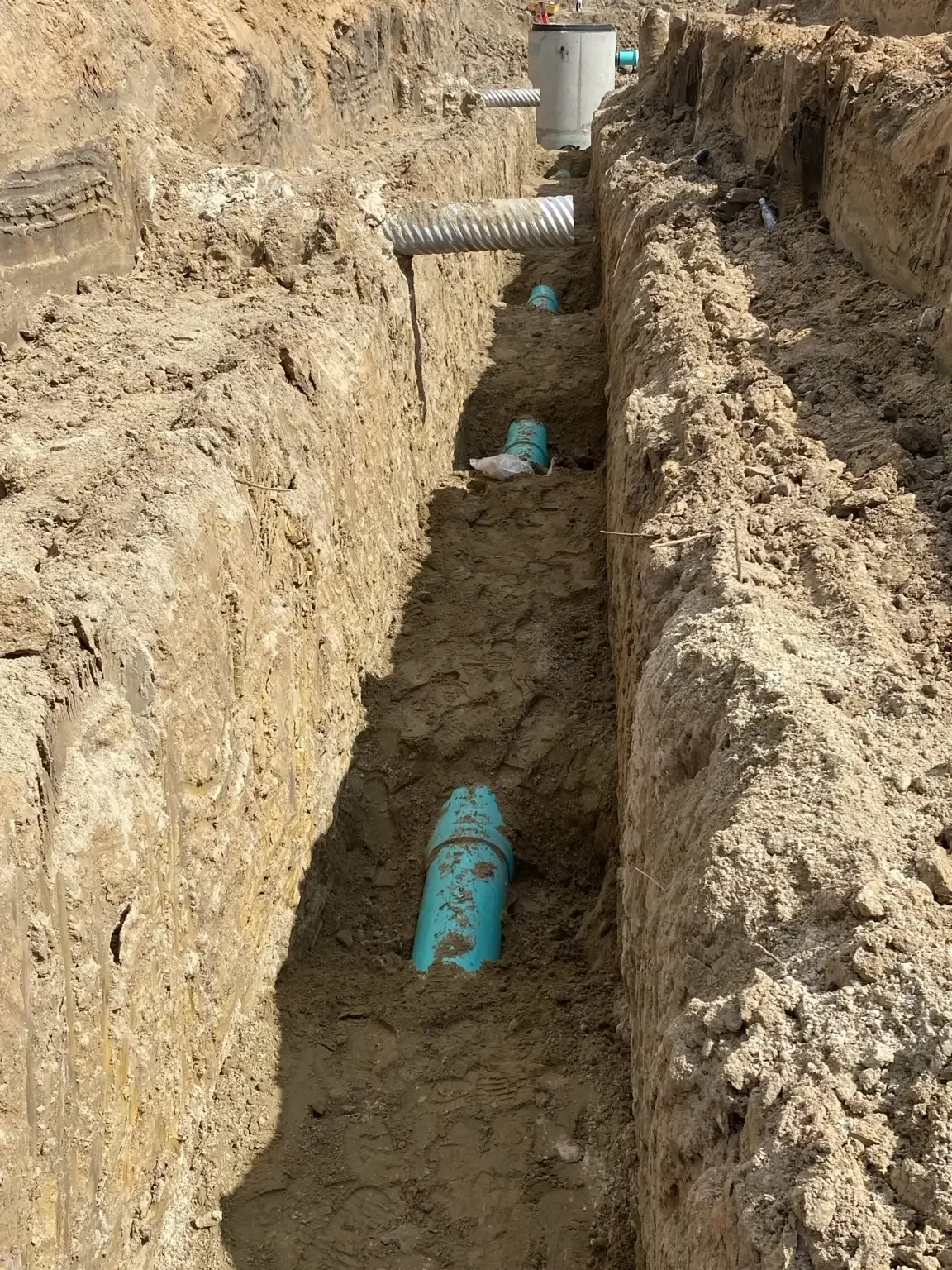View down a long deep utility trench with green PVC sewer pipe segments and crossing utility lines