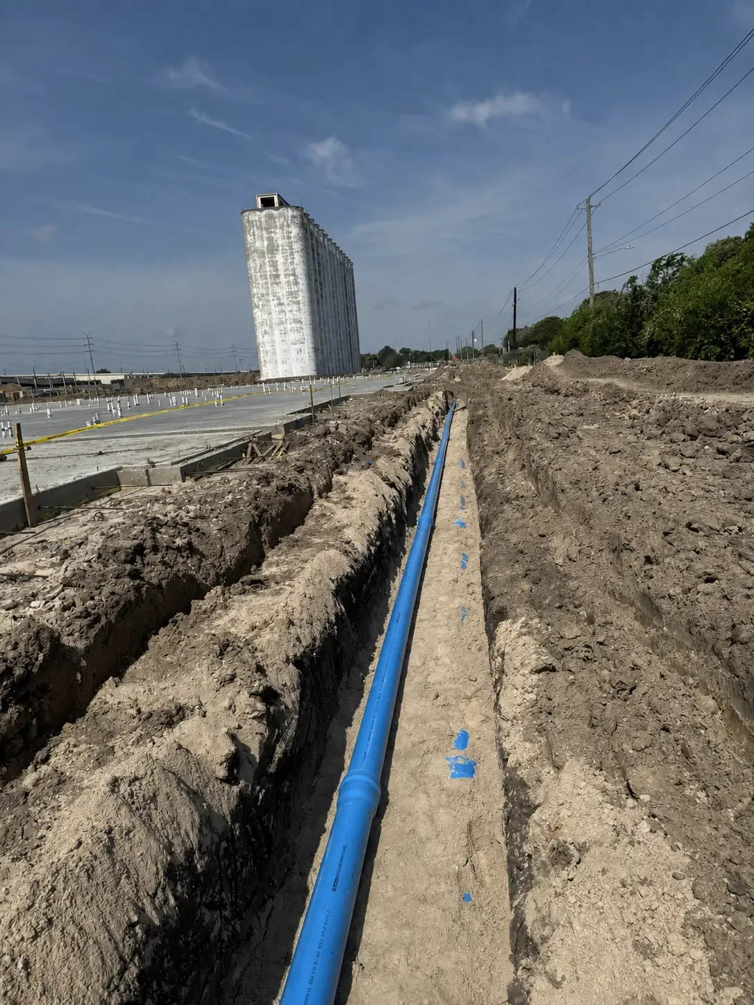 Long straight run of blue PVC waterline installed in a narrow dirt trench alongside a large concrete slab